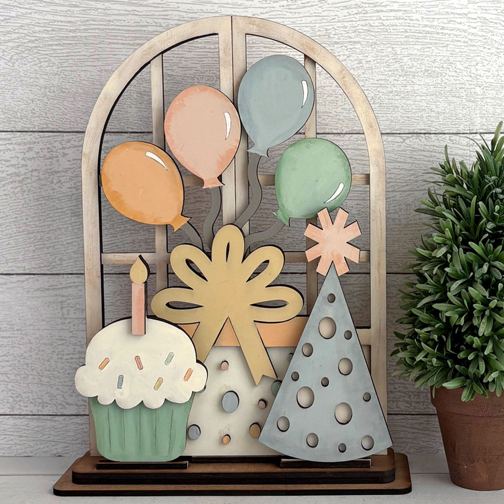Decorative insert for arched doorway with balloons, cake, and tree on a wooden stand against a light wood panel background.