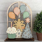 Decorative insert for arched doorway with balloons, cake, and tree on a wooden stand against a light wood panel background.