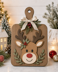 A rectangular wooden cutting board with rounded corners and a handle, decorated with a layered wooden reindeer face, green leafy garlands, red berries, and a cream ribbon bow at the top. The board is photographed on a marble-like surface with holiday decorations, including a miniature house, pinecones, ornaments, and soft string lights in the background.