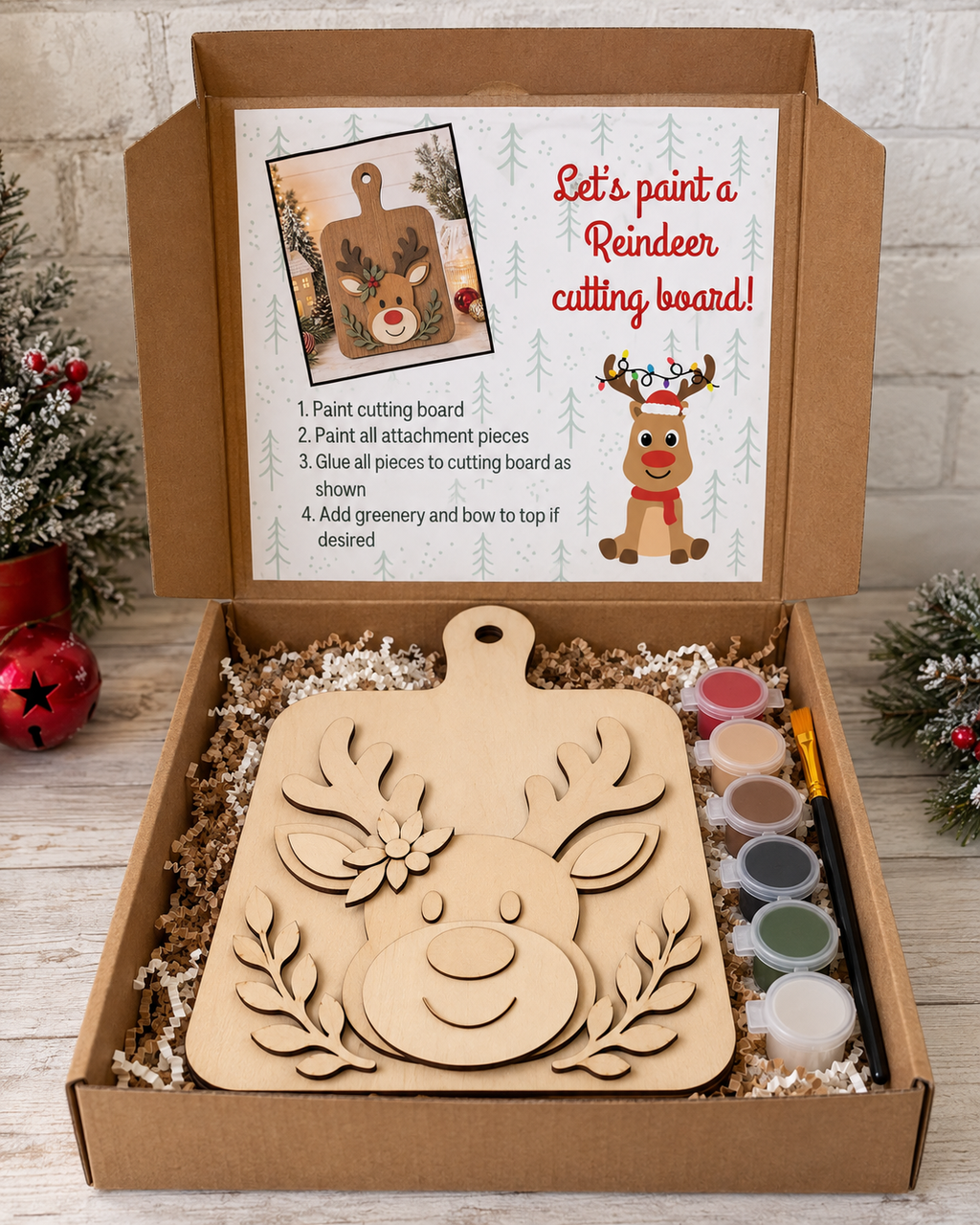 An open cardboard gift box containing a Christmas cutting board kit. The kit includes a large unpainted wooden cutting board with a reindeer face design, a printed instruction card in the lid, six small paint pots, and a pencil, all arranged in crinkle paper filler.