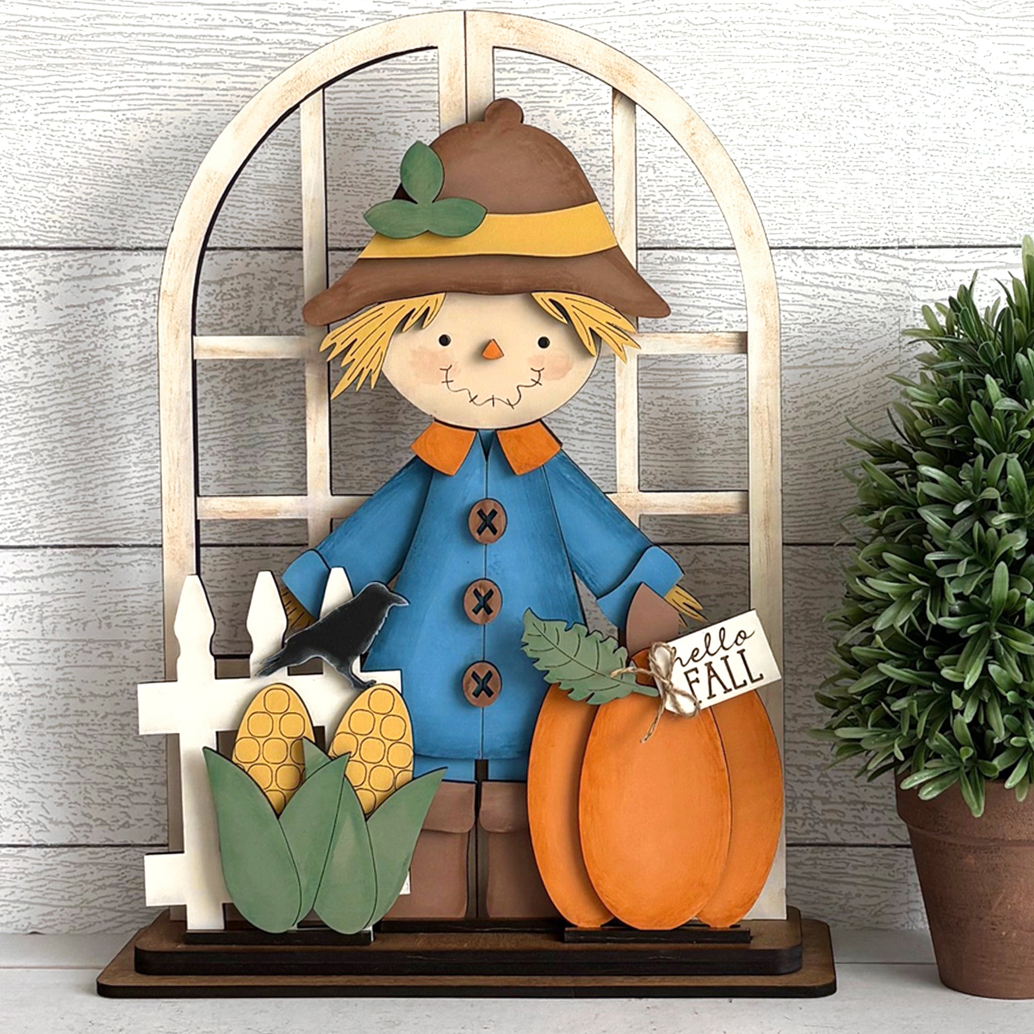 Decorative scarecrow with pumpkins and corn on a wooden stand against a wooden wall.