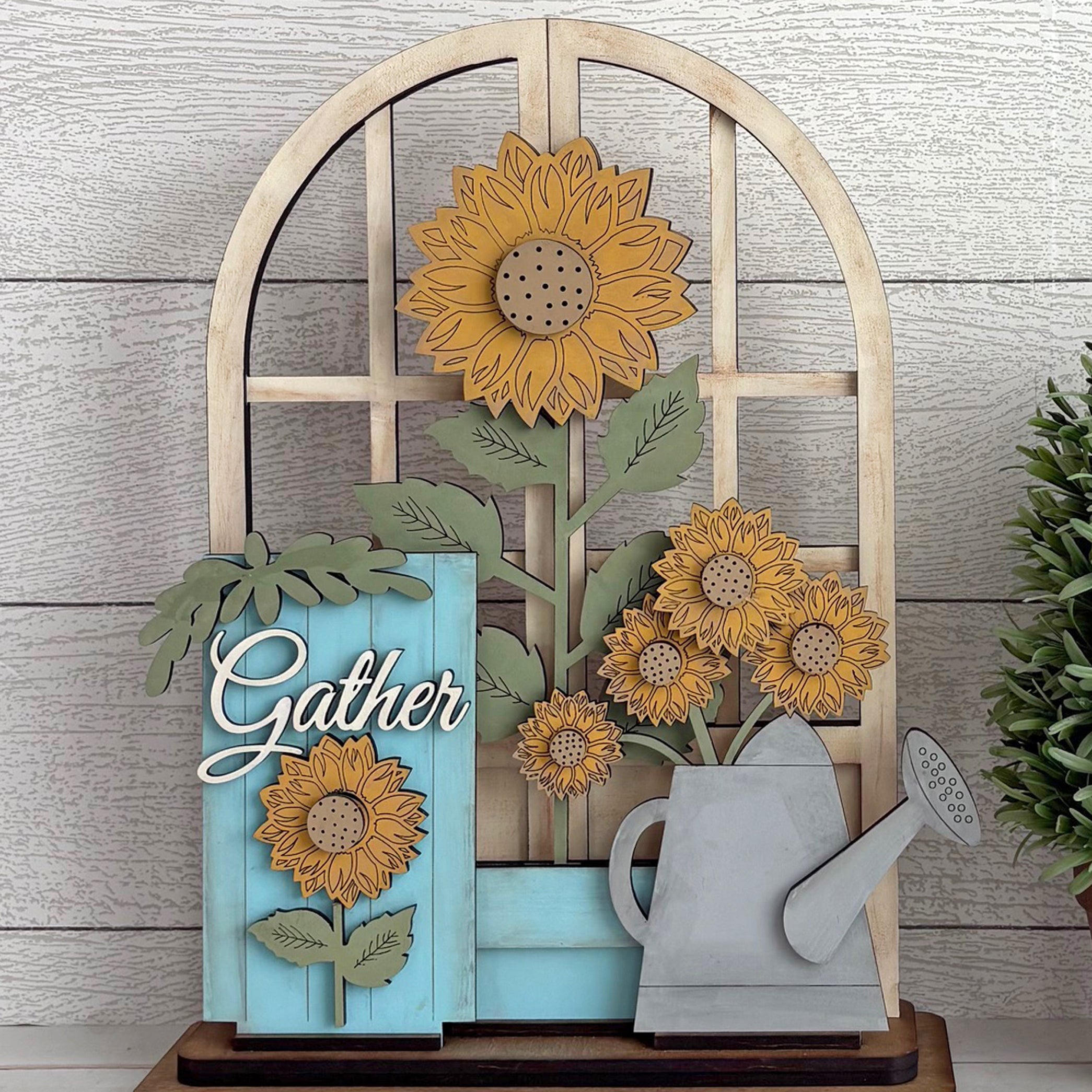 Decorative wall art with sunflowers, a watering can, and 'Gather' sign on a wooden background.