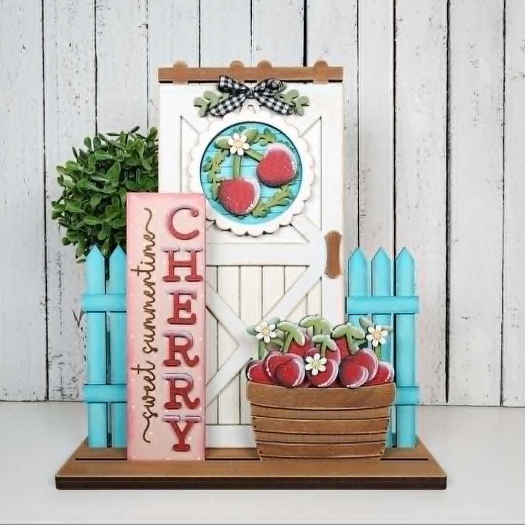 Decorative set with cherry-themed elements including a door, basket, and sign on a white wooden background.